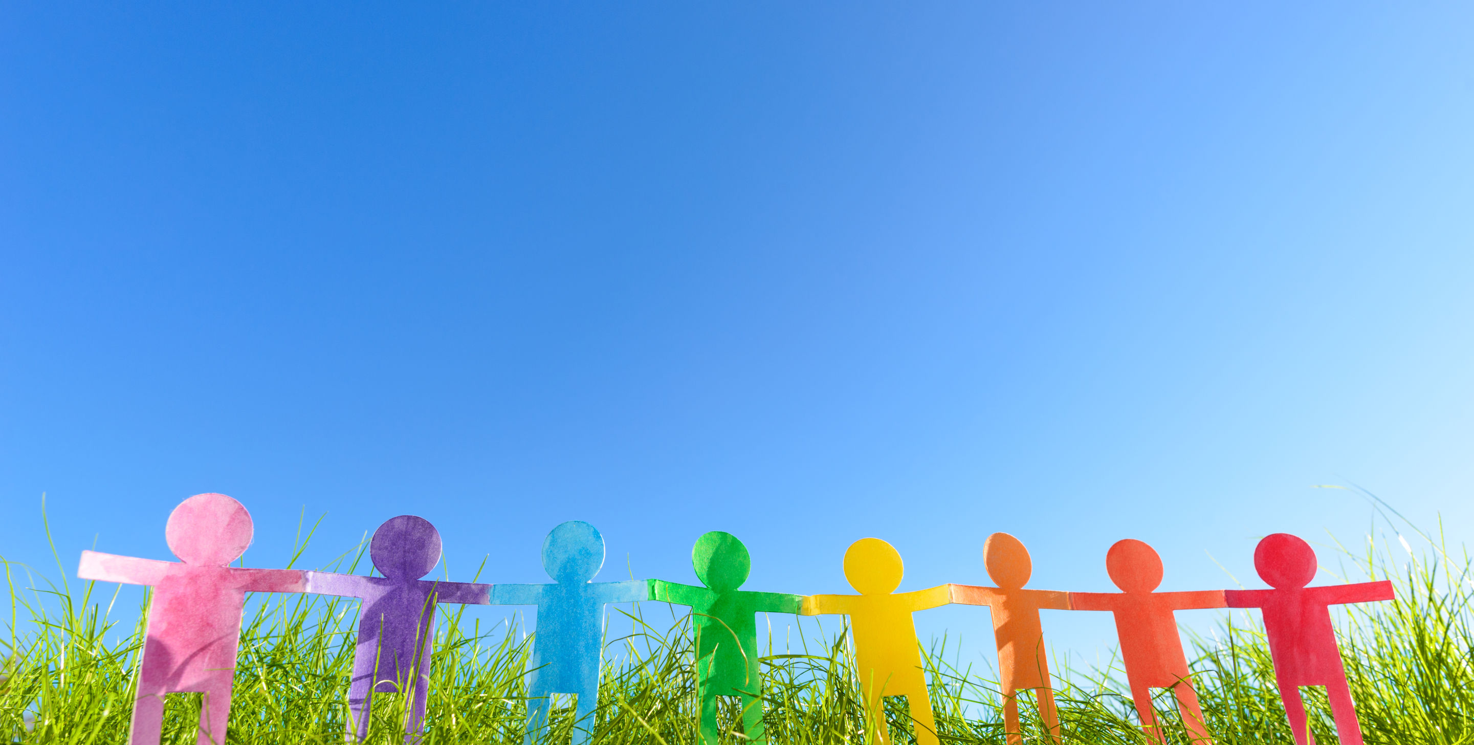 Paper cutouts of faceless mini dolls in different colours, joining hands and standing side by side in a grassland.